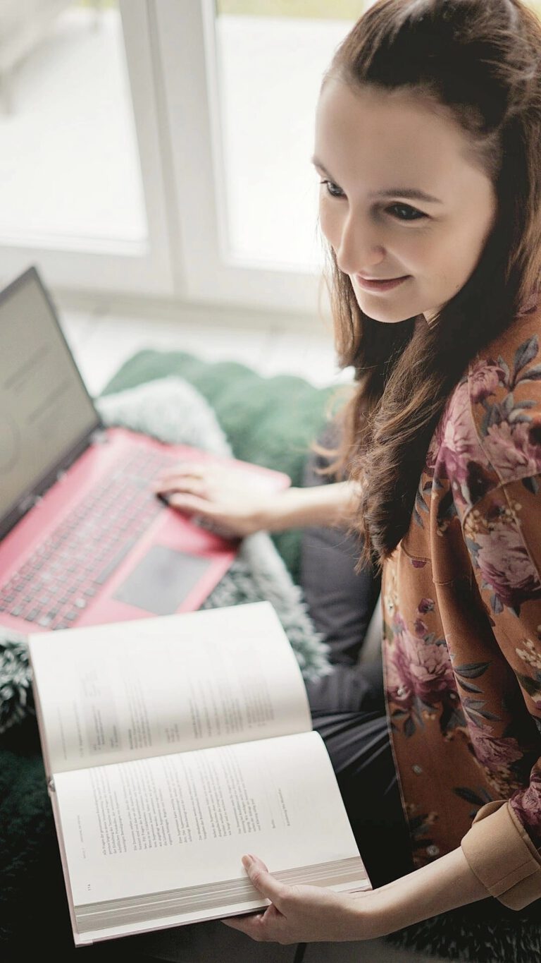 Eine Frau sitzt entspannt am Fenster mit einem aufgeklappten Buch in der Hand und einem Laptop auf dem Schoß. Sie trägt eine geblümte Bluse und lächelt, während sie zur Seite blickt.