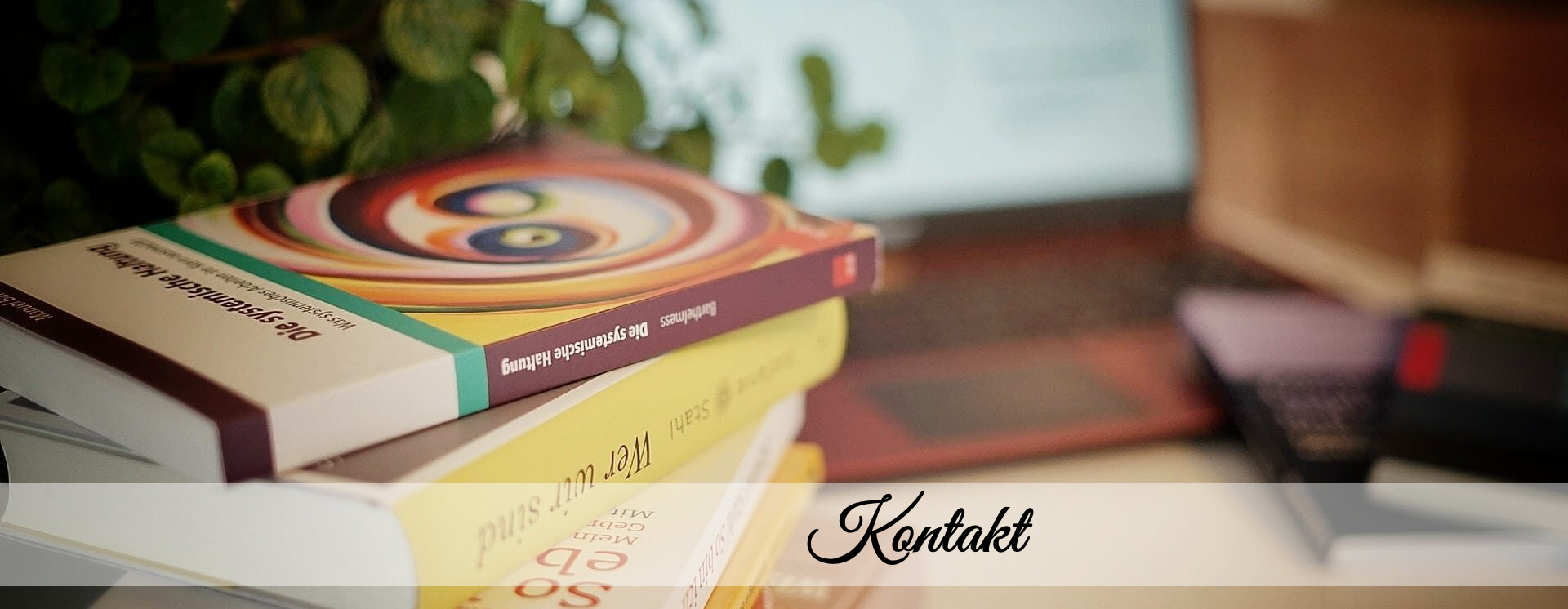 Ein Stapel Fachbücher zu Psychologie und systemischer Beratung auf einem Schreibtisch, im Hintergrund ein Laptop und Notizbücher – im Vordergrund der Schriftzug „Kontakt“.