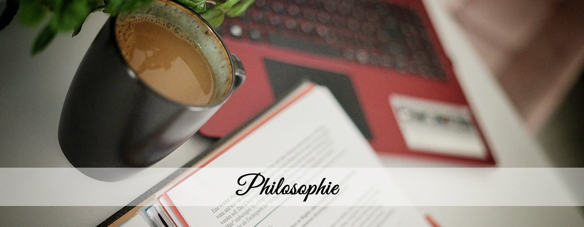 Nahaufnahme eines Arbeitsplatzes mit einer Tasse Kaffee, geöffnetem Buch oder Ordner, rotem Laptop und einer Pflanze – in der Bildmitte der Schriftzug „Philosophie“.