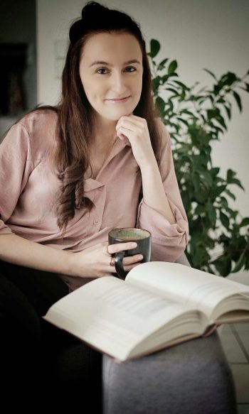 Frau mit langen braunen Haaren sitzt entspannt in einem modernen Wohnraum, hält eine Tasse in der Hand und lächelt in die Kamera, während vor ihr ein geöffnetes Buch liegt.